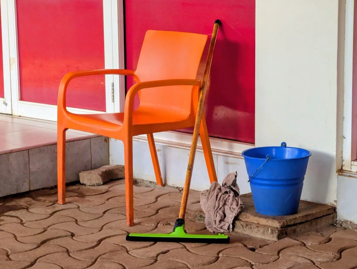 ممسحة أرضيات مطاطية خضراء بمقبض خشبي وقطعة قماش ودلو أزرق بجوار كرسي بلاستيكي برتقالي